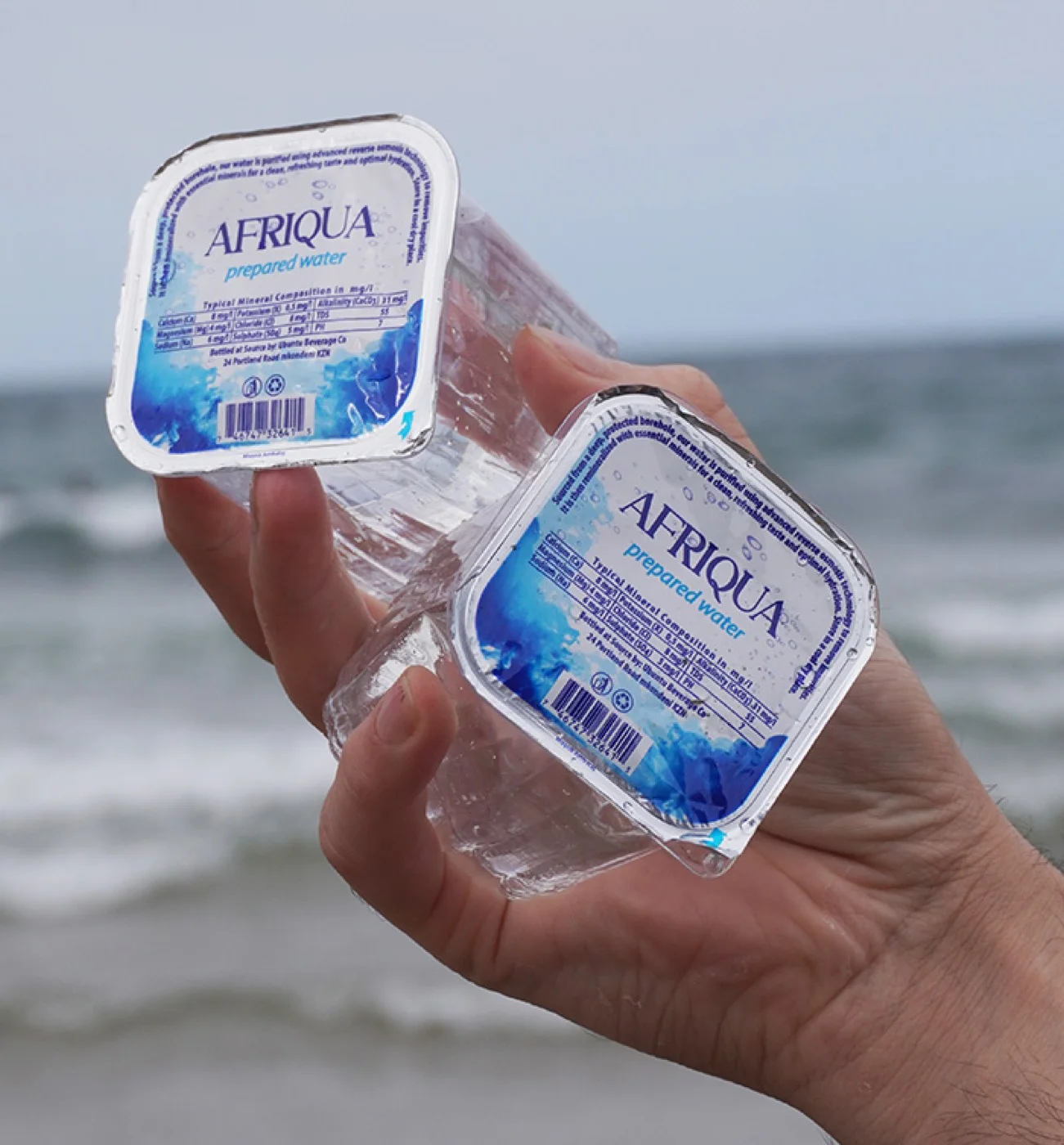 A hand holding two foil-sealed Afriqua prepared water cups at the beach